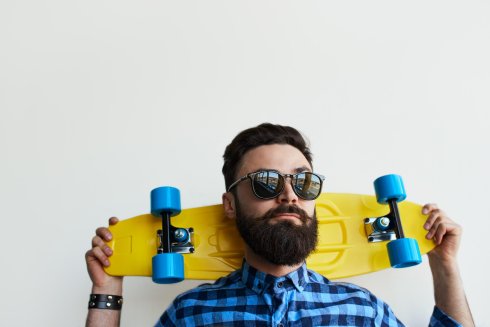 Mann mit Bart und Sonnenbrille hält ein gelbes Skateboard mit blauen Rädern hinter seinem Nacken und steht vor einem schlichten weißen Hintergrund.