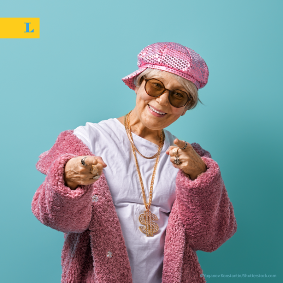 Eine ältere Frau mit rosa Hut und Mantel, Goldkette und Sonnenbrille zeigt lächelnd in die Kamera vor einem blauen Hintergrund.