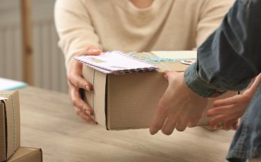 Zwei Personen tauschen über einen Tisch hinweg einen Karton und einen Umschlag aus, wobei nur ihre Hände und Arme sichtbar sind.