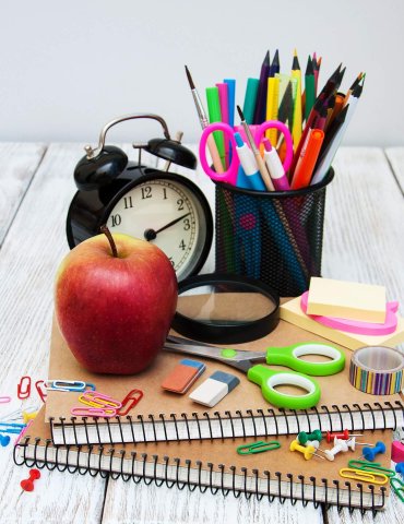 Eine Sammlung von Schulsachen, darunter Notizbücher, ein Apfel, ein Wecker, Stifte, Bleistifte, Scheren, Haftnotizen, Büroklammern und eine Lupe auf einer Holzoberfläche.
