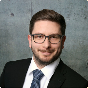 Ein Mann mit Brille, dunklem Anzug, weißem Hemd und gepunkteter Krawatte posiert vor einem grau strukturierten Hintergrund.