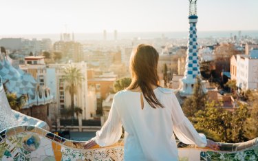 Eine Frau steht auf einem mosaikgefliesten Balkon mit Blick auf das Stadtbild von Barcelona, mit dem Park Güells als markanter Architektur im Hintergrund.