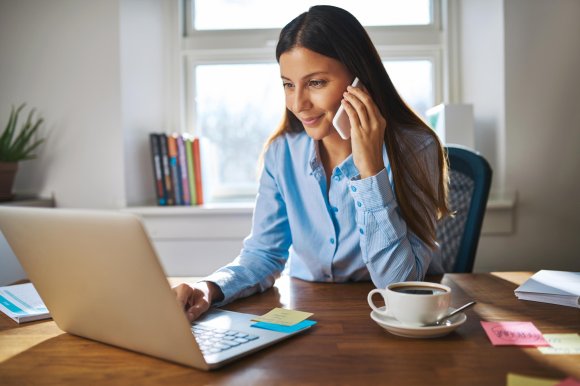 Eine Frau in einem blauen Hemd sitzt an einem Schreibtisch, telefoniert und arbeitet an einem Laptop, in der Nähe stehen eine Tasse Kaffee und Haftnotizen.