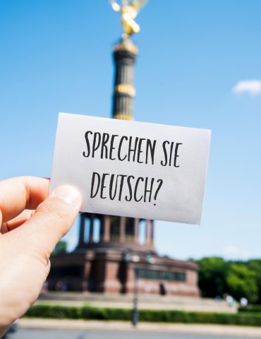 Eine Hand hält eine Karte mit der Aufschrift Sprechen Sie Deutsch? vor der Siegessäule in Berlin vor einem strahlend blauen Himmel.