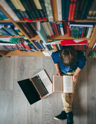 Eine Person sitzt auf dem Boden neben einem Bücherregal und liest ein Buch, daneben steht ein aufgeklappter Laptop.