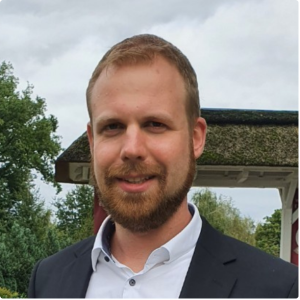 Ein Mann mit Bart und kurzen Haaren, der eine dunkle Anzugsjacke und ein weißes Hemd trägt, steht im Freien vor einem Pavillon und Bäumen.