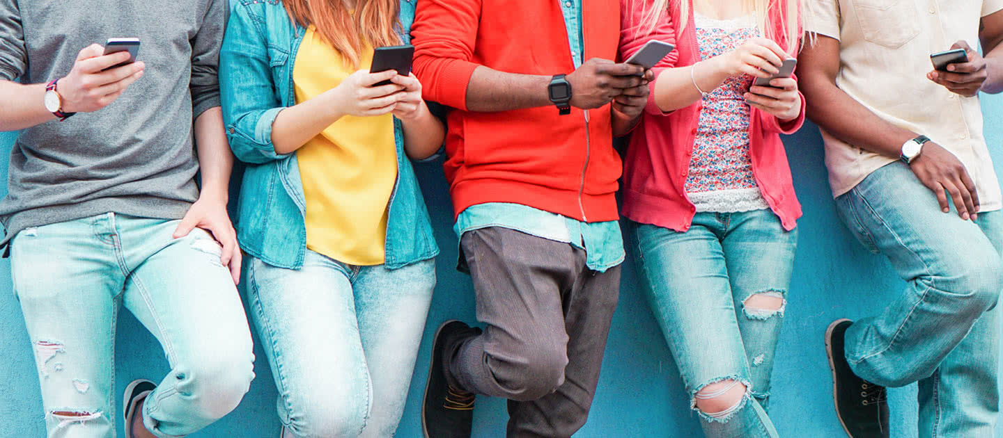 Fünf Personen, die an einer blauen Wand stehen, von den Schultern abwärts, in Freizeitkleidung und mit Smartphones in der Hand.