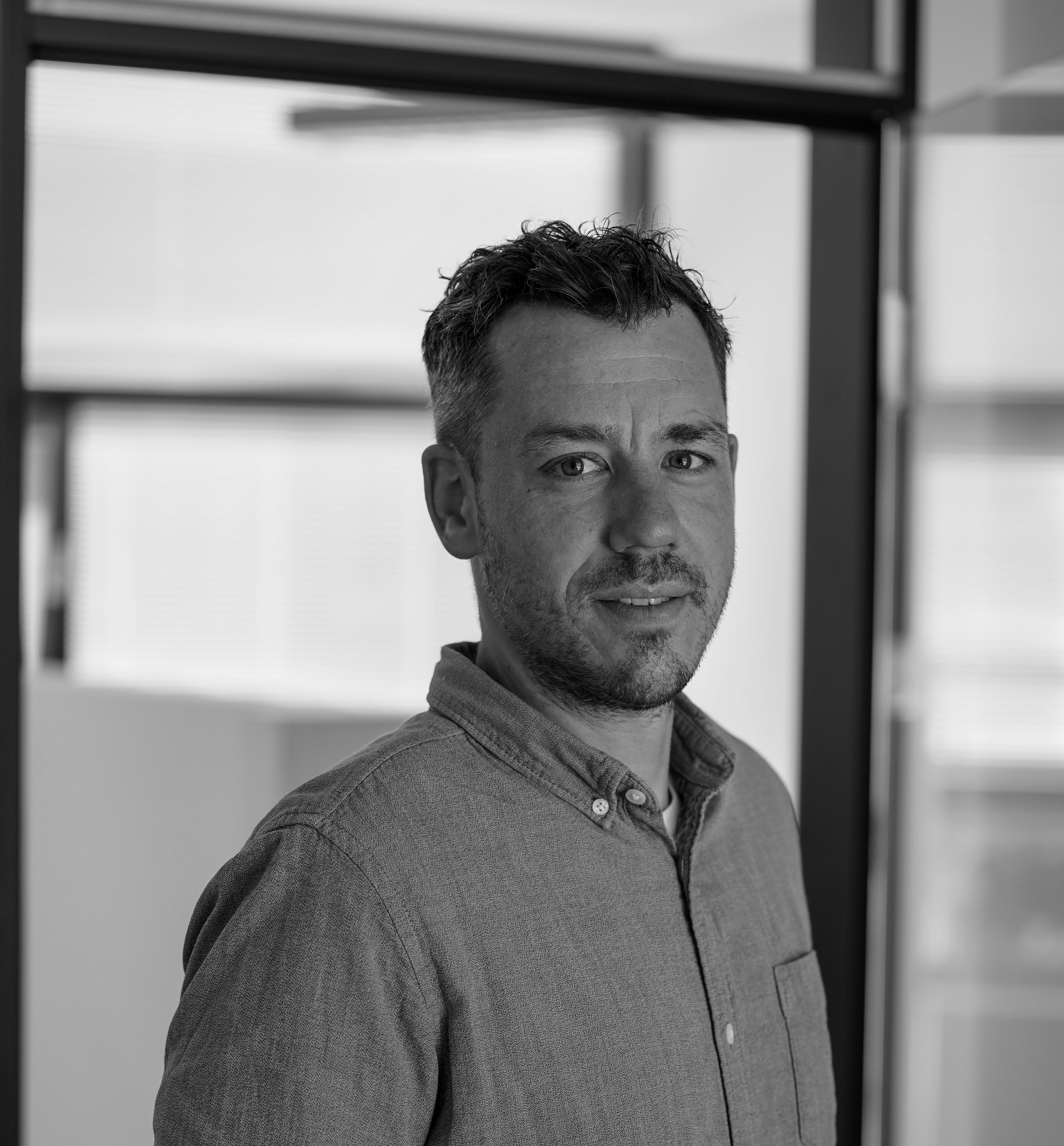 Ein Mann mit kurzen Haaren und Bart steht in einem Innenraum und trägt ein Hemd mit Knöpfen. Das Bild ist in Schwarz-Weiß gehalten, mit Bürofenstern und Glaswänden im Hintergrund.