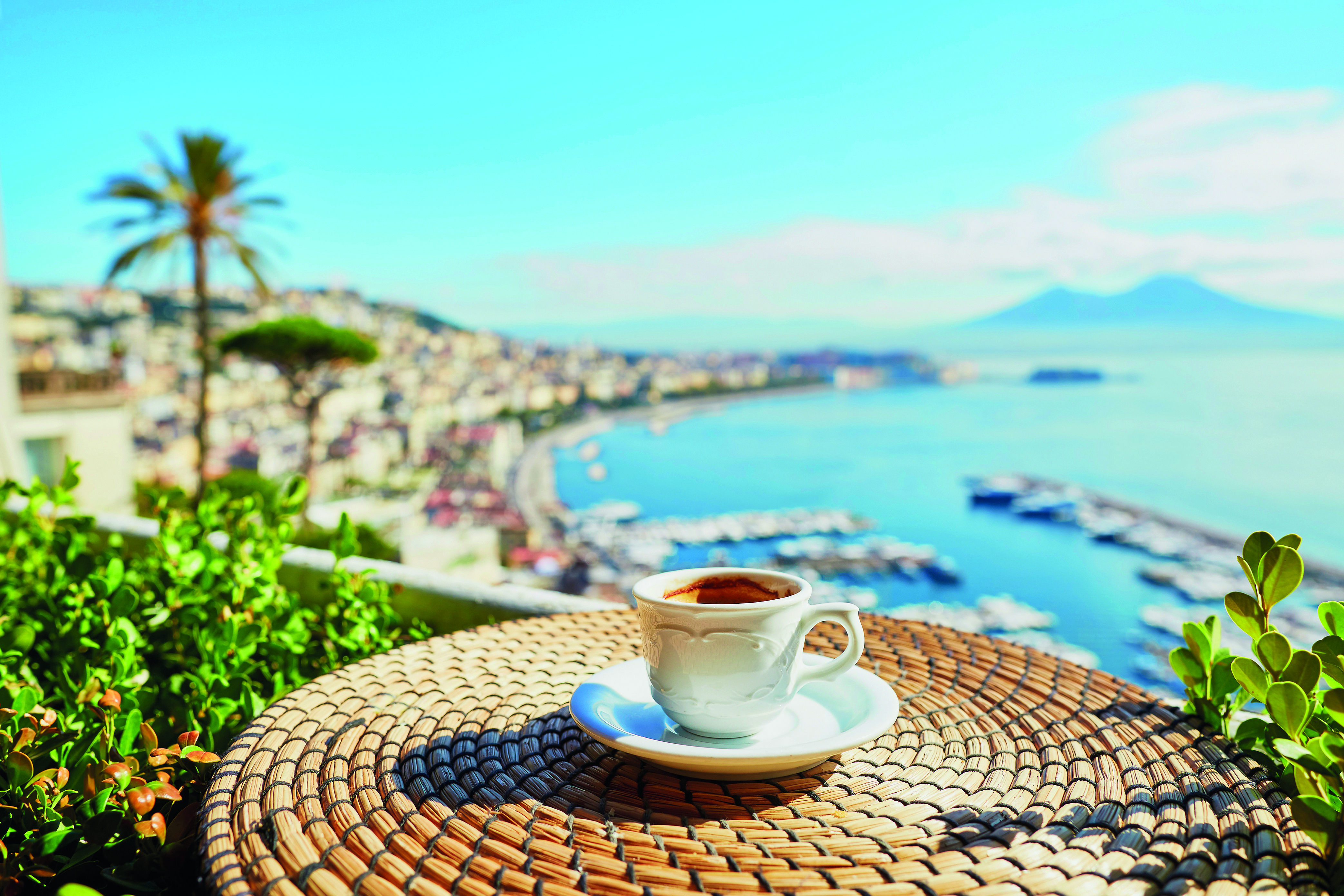 Bei einer Tasse Kaffee auf einem Korbtisch überblickt man eine Küstenstadt, einen Jachthafen und das blaue Meer unter einem klaren Himmel mit fernen Bergen.