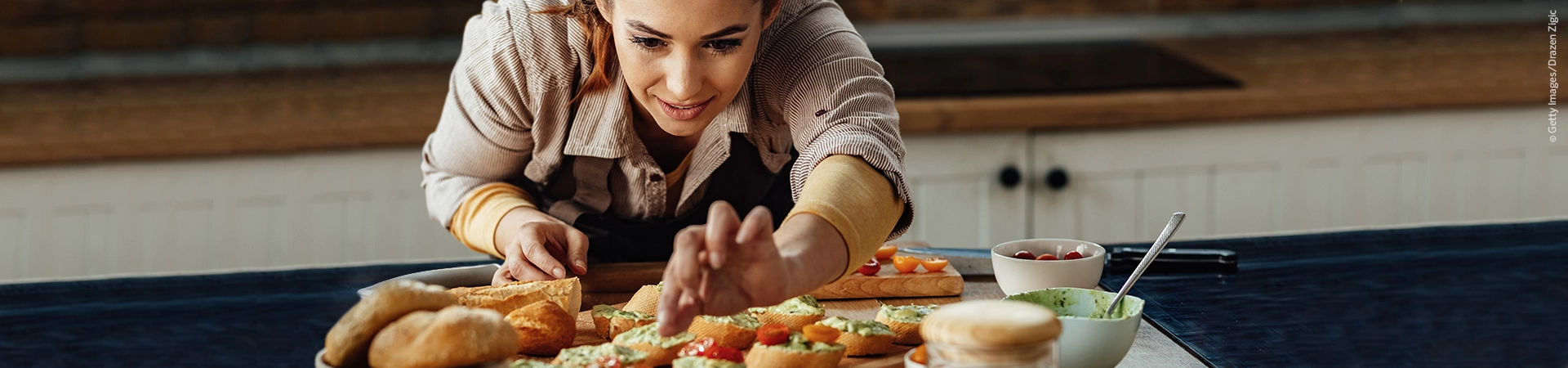 Eine Frau belegt Brotscheiben auf einem Küchentisch, umgeben von Zutaten und Schüsseln, mit Belag.